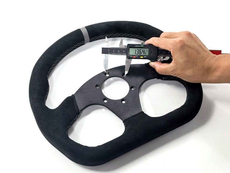 Hand holding a digital caliper to measure a black steering wheel on a white background.