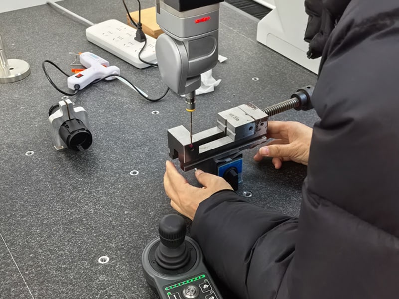 Technician in a dark jacket operating a benchtop drill/milling setup: a metal workpiece is clamped in a vise, with a precision probe above it and a joystick controller in the foreground.