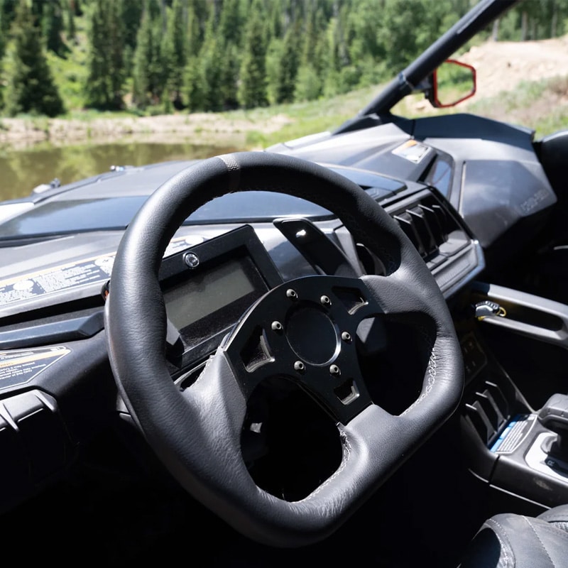 Close-up of a black steering wheel inside an off-road vehicle cockpit with a dashboard, forested scenery visible outside the windshield.