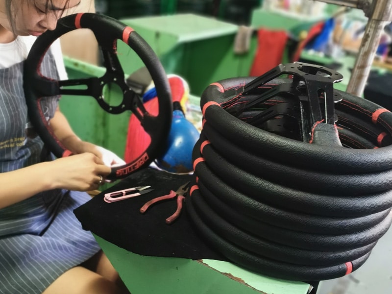 A worker sewing black leather onto a car steering wheel with red stitching at a green workstation, a coil of wrapped steering wheel shells next to her.