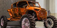 Orange monster truck with oversized tires on display indoors, showcasing its custom build and chrome rims.