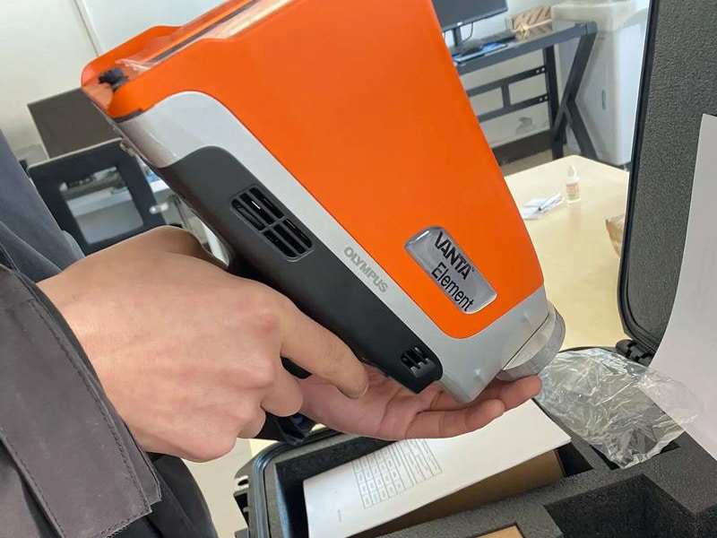 Person holding an orange Olympus Vanta Element handheld XRF analyzer used for material analysis.
