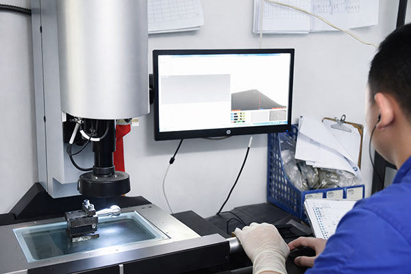 Lab technician operates a benchtop testing instrument with a sample on the stage and a computer monitor on the desk behind.