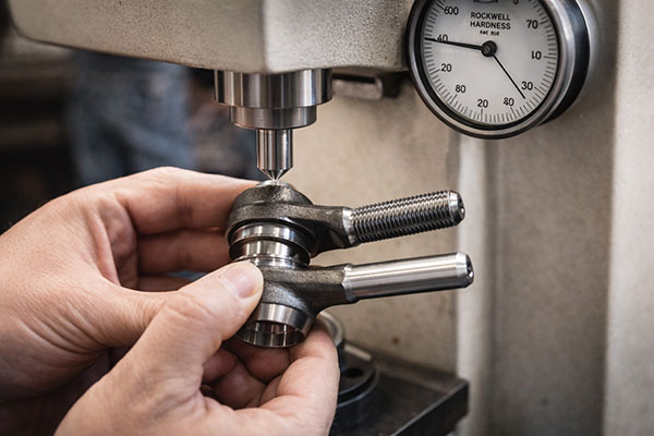 Close-up of a machinist positioning a metal clamp under a drill press spindle, with a hardness gauge in view on the machine side.