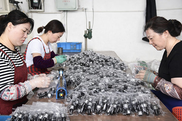 Three workers in a factory packaging small black components into clear bags on a long work table.