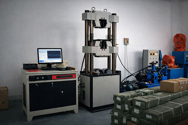 Computer-controlled vertical hydraulic testing machine in a lab, with a monitor on a desk and electrical equipment nearby.