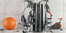 Compact home gym setup featuring a weight-stack machine, exposed pulleys, and an orange exercise ball on a gray floor.