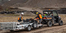 Two workers in orange safety vests and helmets load a quad bike onto a trailer at a dirt construction site, with a utility vehicle nearby.