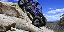Blue off-road vehicle climbing a rocky slope under a bright blue sky, navigating rugged terrain.