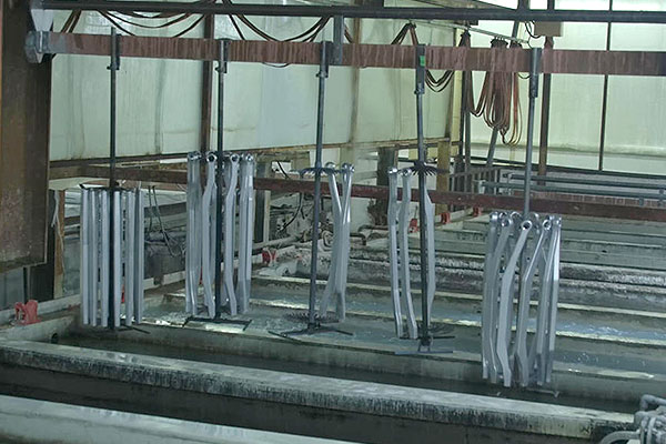 Industrial finishing workshop with multiple curved metal parts hanging from overhead rails, positioned above a large custodial bath/coat tank for coating or cleaning processes.