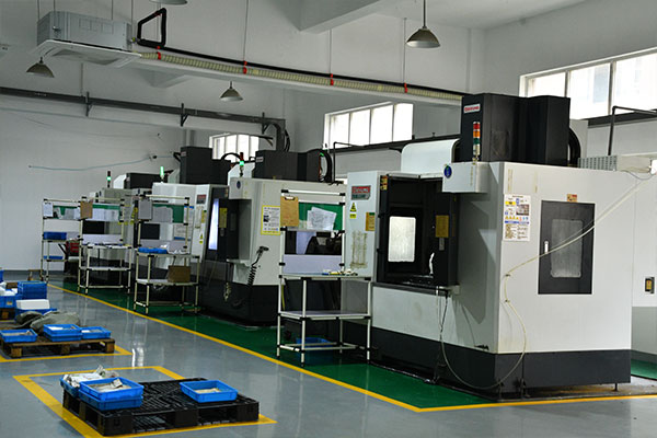 Row of CNC machines in a bright manufacturing workshop, with yellow safety lines on the floor and blue plastic bins on pallets nearby.