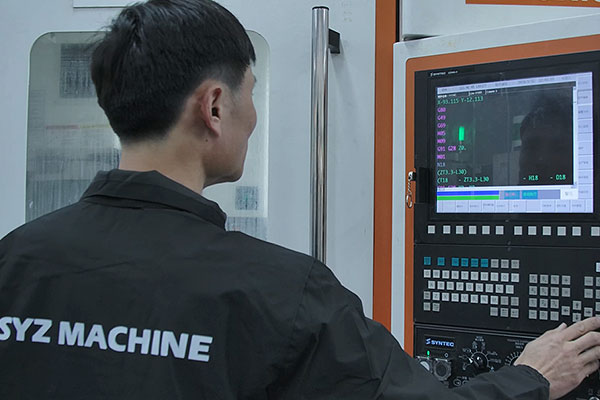 Back view of a technician in a dark jacket with 'SYSZ MACHINE' on the back, operating a CNC machine control panel with a screen displaying numerical data.