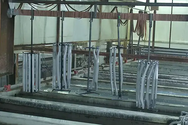 Industrial workshop with multiple metal components hanging from overhead rails, arranged in rows above shallow finishing baths for plating or coating