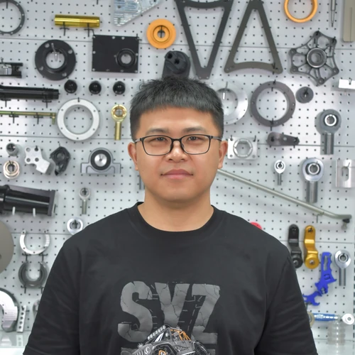 Man with glasses standing in front of a pegboard wall filled with metal tools and parts.