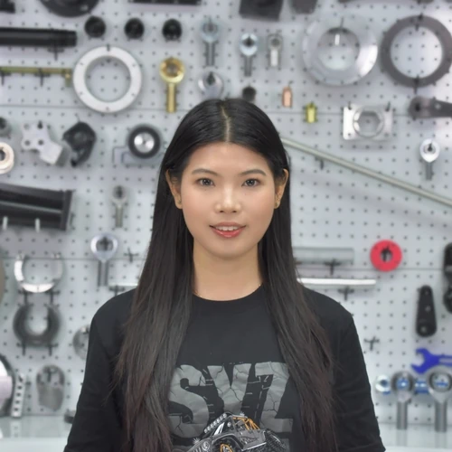 Portrait of a woman with long black hair, standing in front of a wall displaying assorted metal tools and gears.