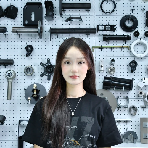 Portrait of a young woman with long dark hair, standing in front of a pegboard wall filled with machine parts in a workshop setting.